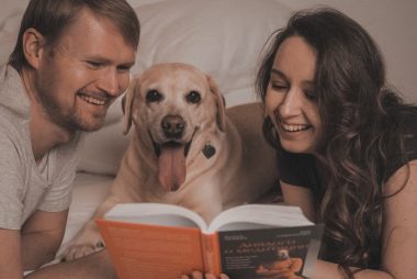 Smiling couple with their dog enjoying a book together in a cozy and relaxed bedroom setting.